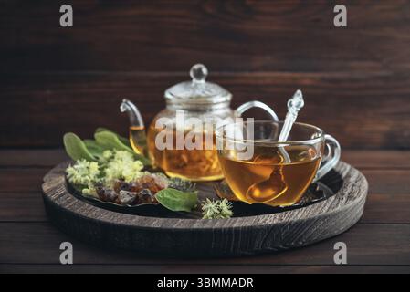 Tazza di tè al tiglio con teiera di vetro, zucchero e fiori di tiglio su vassoio rotondo su sfondo in legno Foto Stock