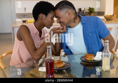 Coppia afroamericana che condivide uno spuntino al tavolo da pranzo della cucina, con piatti e bottiglie Foto Stock