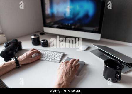 Giovane fotografo freelance che lavora al computer. Progetto artistico creativo. Scrivania con caffè. Tastiera, fotocamera, laptop e obiettivi. Mani maschili io Foto Stock