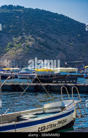Barche attraccate con baldacchini gialli sulle calme acque del porticciolo contro grandi montagne rocciose con vegetazione sparsa sotto il cielo azzurro. Foto Stock