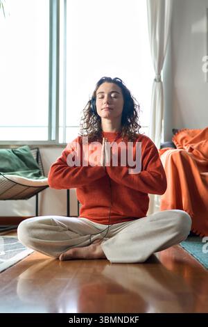 Giovane donna consapevole che indossa cuffie che fanno meditazione a casa. Verticale Foto Stock
