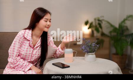 La donna asiatica adulta rilassata pratica l'aromaterapia per alleviare lo stress utilizzando un diffusore di olio essenziale a casa. Promuove il benessere olistico la vita pacifica Foto Stock