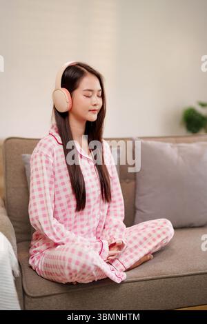 La giovane donna asiatica serena trova calma nella meditazione con il paesaggio sonoro attraverso le cuffie sul divano, promuovendo il benessere olistico e la pace a casa Foto Stock