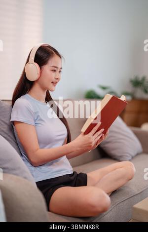 La giovane donna asiatica si rilassa su un comodo divano leggendo un libro indossando le cuffie per creare un'atmosfera tranquilla e consapevole Foto Stock