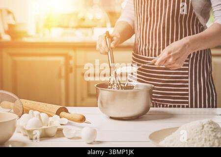 Giovane donna tenendo la coppa con impasto e mescolare con una frusta, primo piano. Una donna in un grembiule a strisce è la cucina in cucina Foto Stock