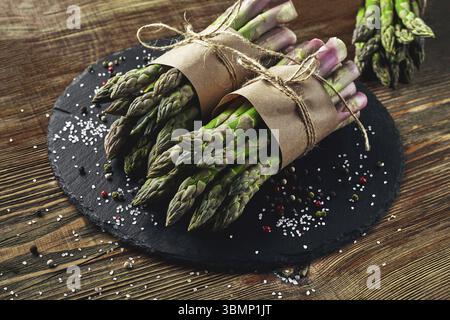 Mazzi di gambi di asparagi commestibili su un'ardesia di pietra, fondo in legno. Verdure fresche e verdi con condimenti, vista dall'alto. Sano me Foto Stock