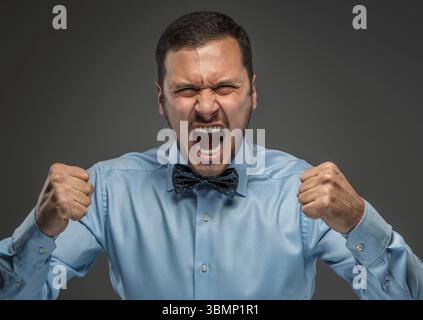 Ritratto di un giovane arrabbiato con camicia blu e cravatta a farfalla con pugni urlanti isolato su sfondo grigio studio. Emozione umana negativa, fa Foto Stock
