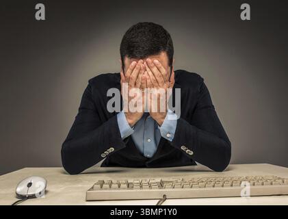 Ritratti un giovane frustrato che copre il viso con le mani mentre è seduto a una scrivania vicino a un computer, isolato su sfondo grigio. Sentirsi male e stanco - Foto Stock