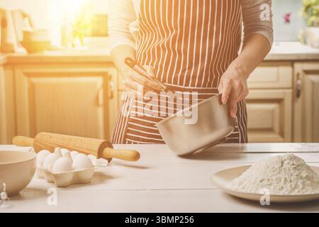 Giovane donna tenendo la coppa con impasto e mescolare con una frusta, primo piano. Una donna in un grembiule a strisce è la cucina in cucina Foto Stock