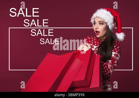 Donna di natale. Modello di bellezza ragazza in santa hat su sfondo rosso. Divertente ridere sorpreso ritratto di donna. A bocca aperta. Emozioni vere. Labbra rosse e essere Foto Stock
