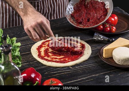 Un maschio di spargimento a mano la salsa di pomodoro sulla base per pizza con cucchiaio su un vecchio sfondo di legno. Concetto di cucina a vista. Close-up Foto Stock