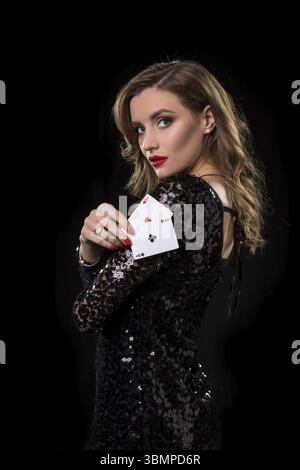 Giovane bella donna in abito nero lucido tiene le carte da poker in mano su uno sfondo nero. Il concetto di gioco d'azzardo. Casinò Foto Stock