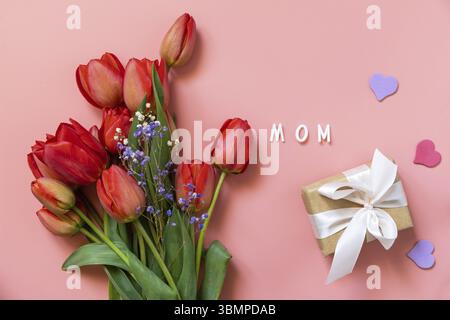 Tulipani rossi con scatola regalo e cuori piccoli, testo scritto a mano dalla mamma su sfondo rosa pastello con spazio vuoto. Concetto per la festa della mamma. Visualizza Foto Stock