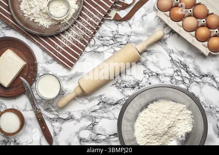 Primo piano. Vista dall'alto degli ingredienti da forno e degli utensili da cucina sullo sfondo del tavolo in marmo. Ci sono una serie di uova, farina su un piatto e in un fiocco Foto Stock