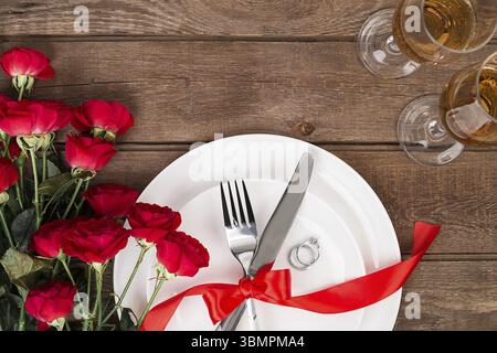 Cena di San Valentino messa in tavola con nastro rosso, rose, coltello e forchetta e anello su sfondo di quercia. Still Life Foto Stock