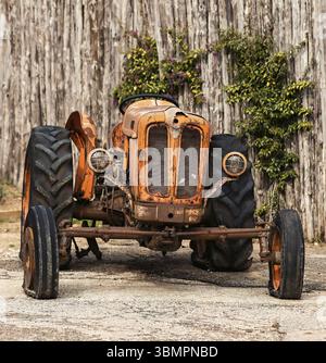 Il vecchio trattore arancione arrugginito è parcheggiato di fronte a una recinzione di legno in un'azienda agricola Foto Stock