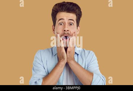 Ritratto di un giovane uomo spaventato con una camicia blu su sfondo beige che guarda la fotocamera. Foto Stock