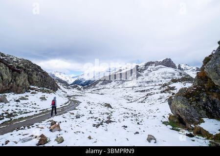 l'escursionista da solo irriconoscibile con uno zaino rosso cammina lungo un sentiero innevato circondato da cime rocciose sotto un cielo nuvoloso Foto Stock