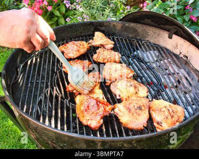 Spennellare le cosce di pollo con una marinata al barbecue appiccicosa in una calda serata estiva in un cortile con carboni caldi Foto Stock