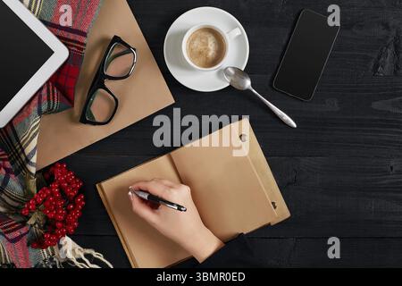 Giovane donna destra scrivere su notebook vuoto su tavolo di legno con caffè tazza, smartphone, e bicchieri accanto al mattino tempo. Vista dall'alto. Vita morta. Foto Stock