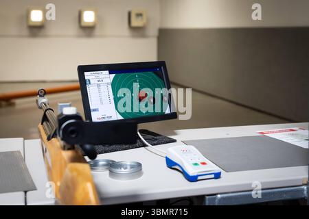 Landkreis Osnabrück, Deutschland 25. Juni 2025: IM Bild: Eine breitere Ansicht eines modernen Schießstands zeigt die Kombination aus traditionellem Sportgerät und fortschrittlicher Technologie. Im Vordergrund liegt ein klassisches Luftgewehr mit Holzschaft, bereit für den nächsten Schuss. Daneben befinden sich zwei Dosen mit Diabolos und eine weiße Fernbedienung mit blauen und roten Tasten zur Steuerung des Systems. Monitor LCD IM Zentrum des Bildes steht ein, der die elektronische Zielscheibe und die hervorragenden Ergebnisse des Luftgewehr LG Auflage 30 Schießens anzeigt, mit einer Gesamtpun Foto Stock
