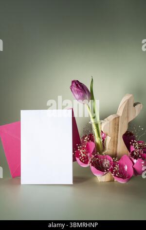 Modello di carta bianca vuota mock up cartolina decorazione per le festività pasquali con materiali sostenibili guscio d'uovo color rosa decorazioni in vaso coniglietto in legno con Foto Stock