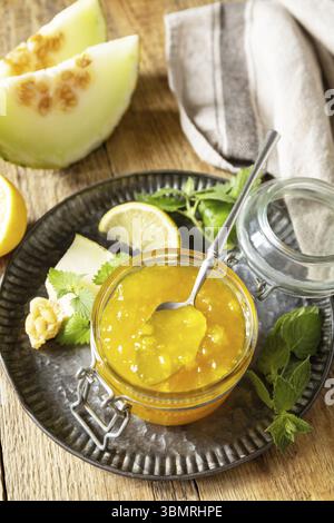 Riserva naturale fatta in casa. Confettura dolce di melone e agrumi o gelatina in un piccolo vaso di vetro con fette di melone fresche su un tavolo rustico in legno Foto Stock