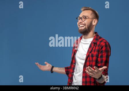 Emotivo e persone concetto: giovane uomo barbuto in camicia a scacchi. Hipster stile. Studio shot su sfondo blu. Spazio di copia Foto Stock