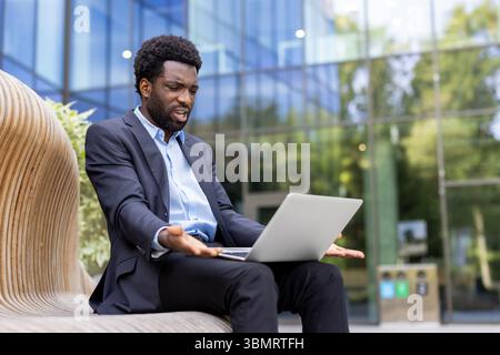 Un uomo d'affari frustrato con una tuta reagisce mentre guarda il suo notebook. All'aperto, vicino ad un edificio moderno. Foto Stock