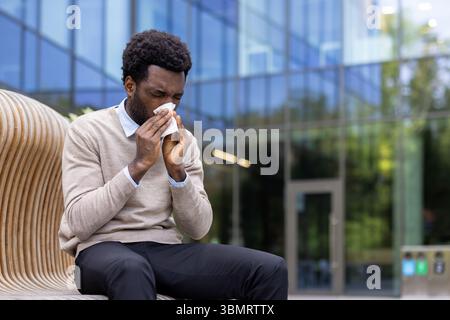 Un afroamericano soffia il naso mentre si siede all'aperto di fronte a un edificio moderno. Foto Stock
