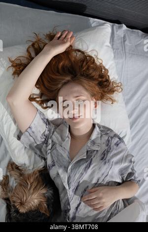 Una giovane donna dorme tranquillamente nel suo comodo letto con il suo amato cane, circondato da morbide e confortevoli lenzuola Foto Stock