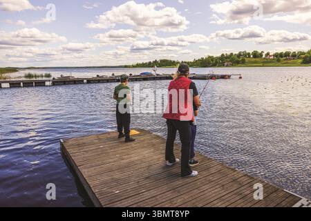 Madre che pesca con due bambini dal molo di legno sul lago durante le soleggiate giornate estive. Svezia. Foto Stock