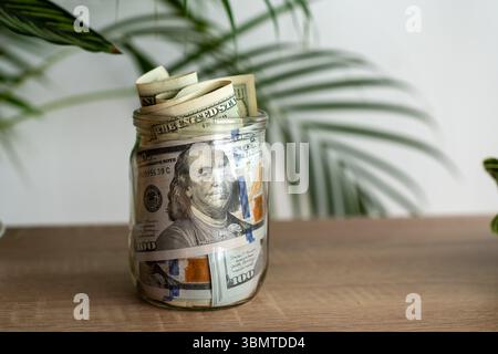 Barattolo di vetro pieno di banconote da cento dollari arrotolate e impilate su un tavolo di legno con piante tropicali verdi sullo sfondo. Foto Stock