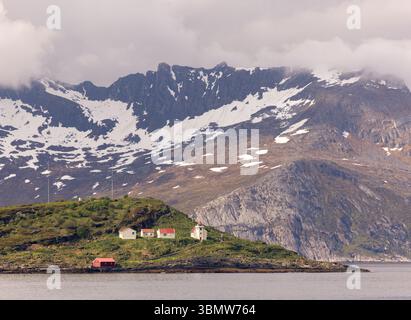 Faro e piccole case su un'isola remota nella Norvegia artica, con spettacolari montagne spolverate di neve alle spalle e condizioni di disgelo primaverile. Foto Stock