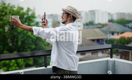Giovane uomo creativo che scatta una foto della sua tazza da caffè, ammirando la vista dal balcone della città Foto Stock
