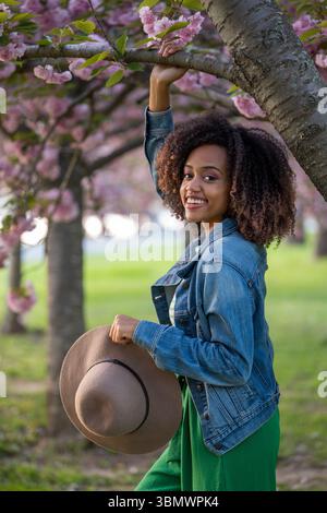 Una giovane donna afroamericana con capelli naturalmente ricci tiene un cappello di paglia nella mano sinistra. Foto Stock