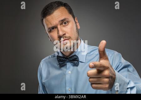 Il ritratto ravvicinato di un uomo con la camicia blu e la cravatta a farfalla è un gesto con la mano, puntando il dito verso la fotocamera isolata su sfondo grigio Foto Stock