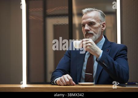 Ritratto di un uomo d'affari anziano che indossa un abbigliamento formale tenendo in mano una tazza di caffè mentre contempla. Uomo dai capelli grigi con barba seduto a un tavolo di legno in un moderno ambiente da ufficio Foto Stock