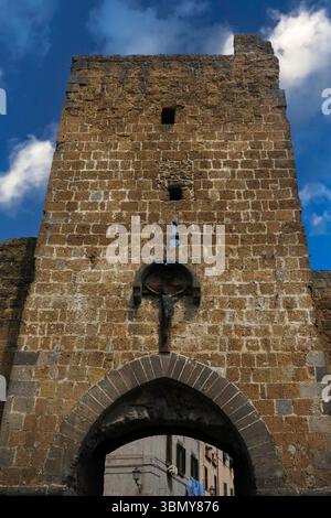 Porta San Marco, usurata dal tempo ma imponente, tra le porte medievali e le torri di guardia nelle mura difensive che proteggevano l'ex città etrusca e romana di Tuscania nella provincia di Viterbo, Lazio, Italia. Costruito in tufo vulcanico dorato e grigio con crocifisso a baldacchino su arco. Foto Stock
