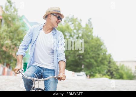 Uomo in bicicletta lungo una strada asfaltata alberata vicino alle pareti di mattoni sotto un cielo limpido e soleggiato Foto Stock