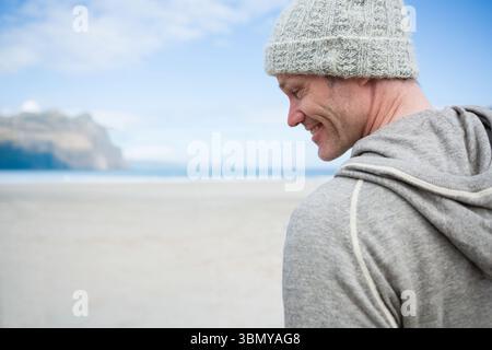 Uomo che indossa un berretto grigio e una felpa con cappuccio in piedi sulla spiaggia sabbiosa e sorride verso le scogliere, copia lo spazio Foto Stock