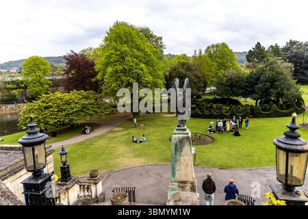 Bath, Regno Unito - 25 aprile 2025 - Vista dei giardini Parade con la statua dell'angelo di bronzo, le persone sul prato e gli edifici storici nel Backgro Foto Stock