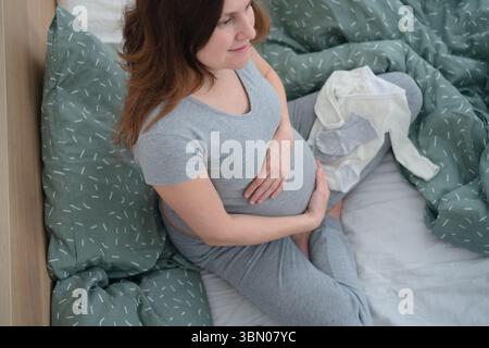 Una donna incinta incinta si siede a gambe incrociate, circondata da vestiti per bambini, toccando delicatamente la pancia. Un momento tranquillo di amore e anticipazione. Foto Stock