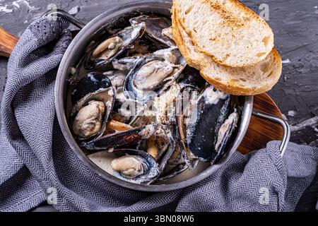 Cozze in conchiglie cotte in una cremosa salsa all'aglio e toast tostato in una casseruola con un tovagliolo grigio, su sfondo grigio. Vista dall'alto. Pol. Di alta qualità Foto Stock