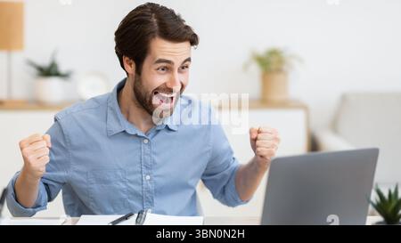 Eccitato giovane uomo d'affari al computer portatile scuotendo i Fists in ufficio Foto Stock