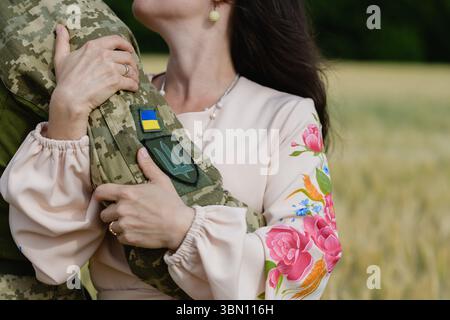 Primo piano di una donna con un tradizionale abito ricamato ucraino che abbraccia un soldato in uniforme mimetica con bandiera e toppa tridente, mostrando emozione Foto Stock