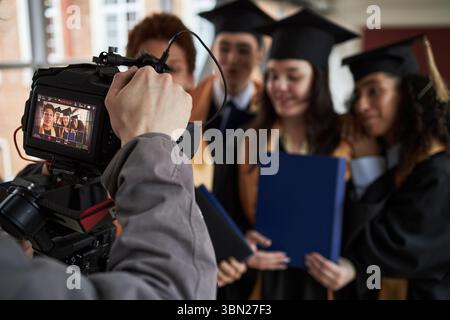 Gruppo di giovani uomini e donne multietnici che indossano berretti e camici che si alzano insieme in posa per la macchina fotografica mentre tengono diplomi durante la cerimonia di laurea universitaria Foto Stock