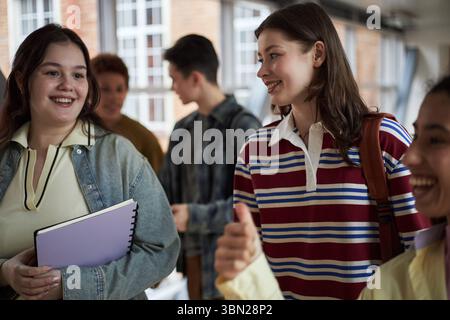 Gruppo di giovani donne e uomini studenti universitari multietnici che camminano insieme in corridoio, sorridenti e chiacchierano, tengono in mano quaderni e zaini, si impegnano in una conversazione amichevole Foto Stock
