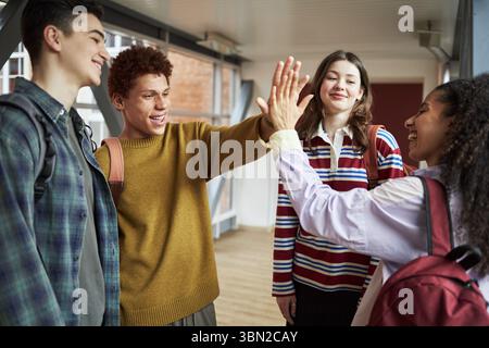 Gruppo di adolescenti multietnici che sorridono e danno alti fiocchi nel corridoio universitario, trasportano zaini e interagiscono tra loro, mostrando un legame sociale positivo tra gli studenti Foto Stock
