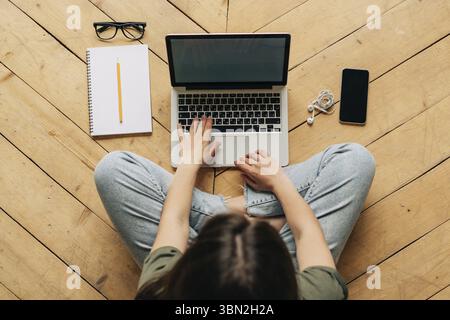 Draufsicht auf eine anonyme Frau, die auf einer Tastatur tippt und mit notebook und Handy auf dem Holzboden sitzt Foto Stock